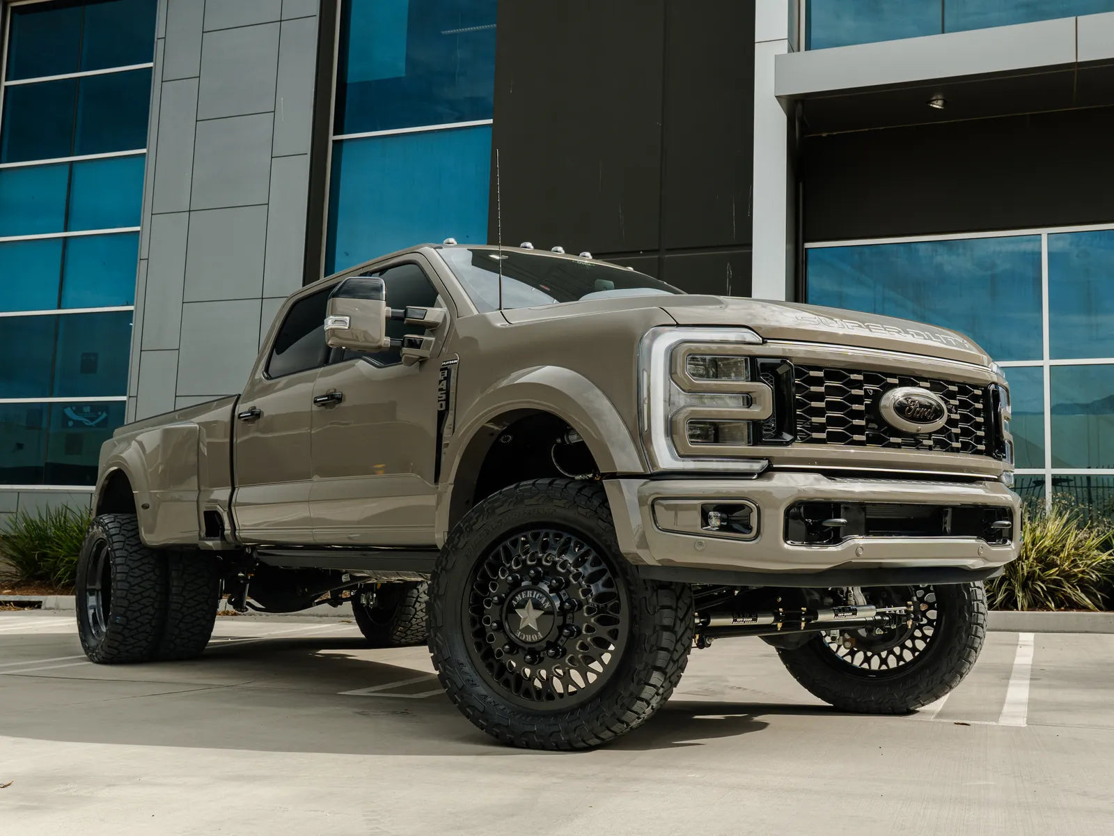 Front three-quarter view of a tan Ford F-450 Platinum dually with black mesh wheels, lift and Ridge Grappler tires in front of a glass building.
