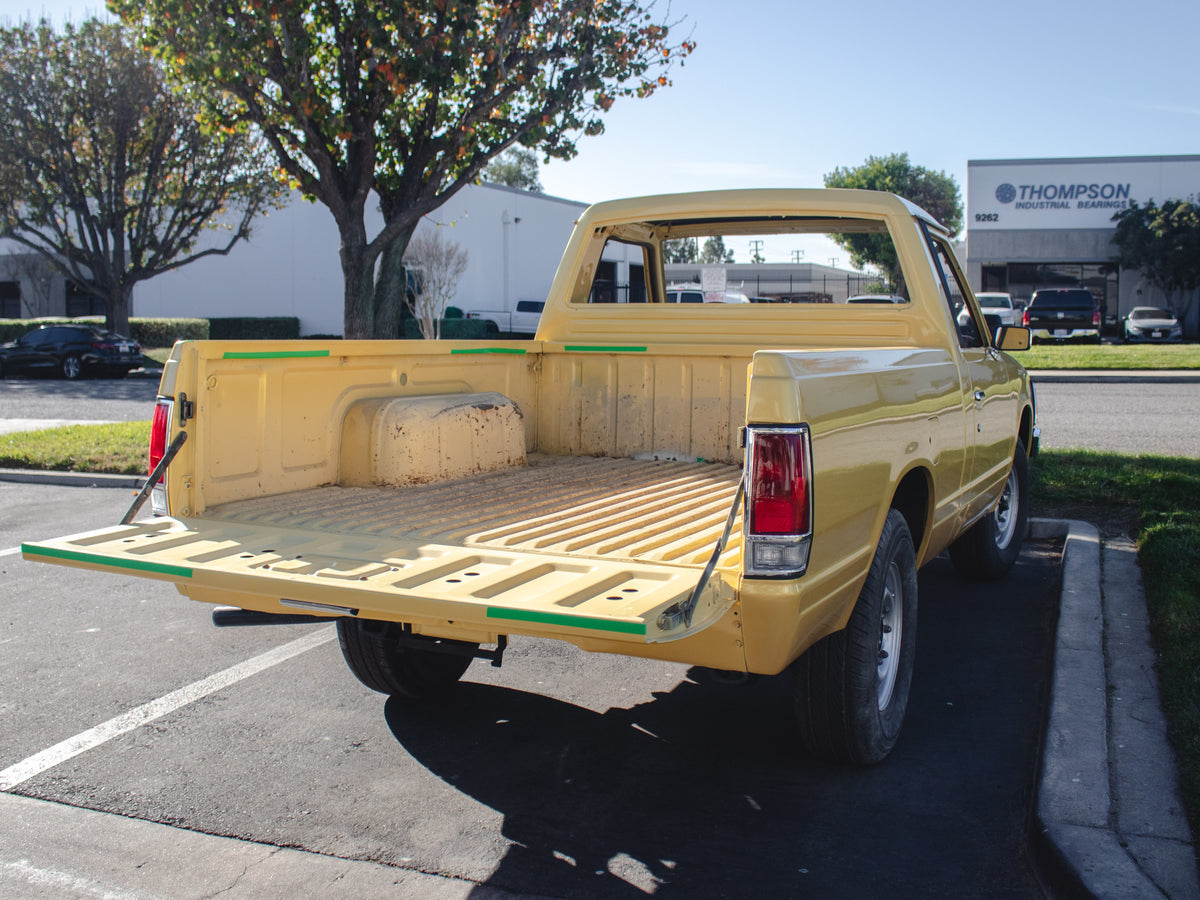 A yellow vintage pickup truck parked in an outdoor lot, viewed from the rear three-quarter angle. The truck bed is empty and shows some wear, with a visible 'T' badge on the tailgate.