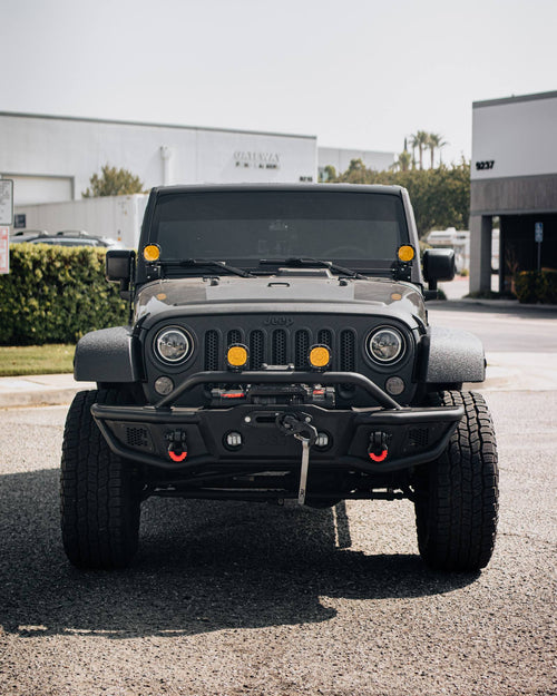 Front view of a black Jeep Wrangler Rubicon with a winch bumper, amber lights and red recovery hooks