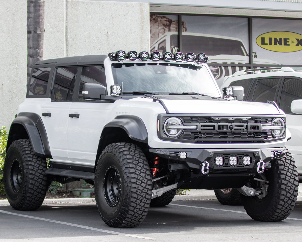 Lifted white Ford Bronco equipped with oversized off-road tires, an ADD front bumper, skid plate, recovery shackles, and roof-mounted auxiliary lights.