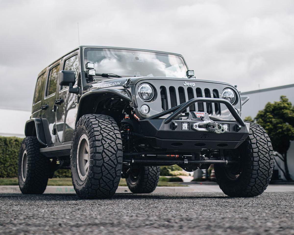 A lifted black Jeep Wrangler with oversized off-road tires, a heavy-duty front bumper, and a winch, photographed outdoors on a paved surface under an overcast sky.