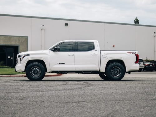 Side profile of a white Toyota Tundra crew cab with black wheels in front of a warehouse