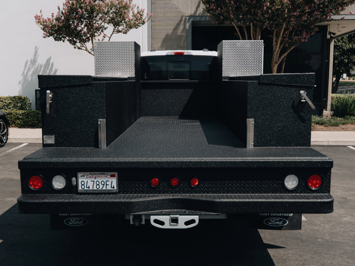 Rear view of a black Ford utility truck bed with diamond plate side walls and a textured black liner, parked outdoors on a paved surface.