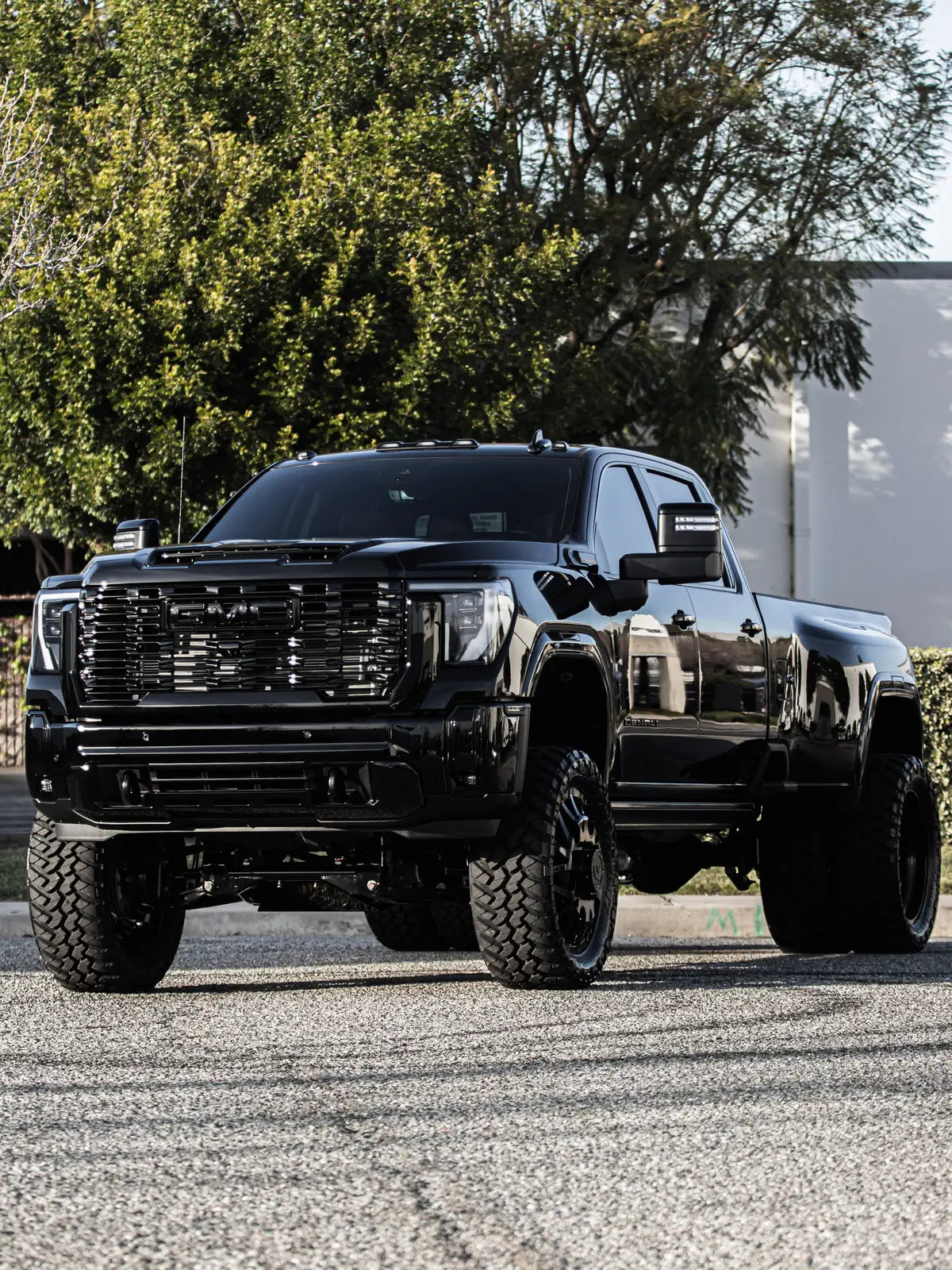 Front view of a glossy black lifted GMC Sierra Denali dually with black wheels and mud tires on a gravel lot.