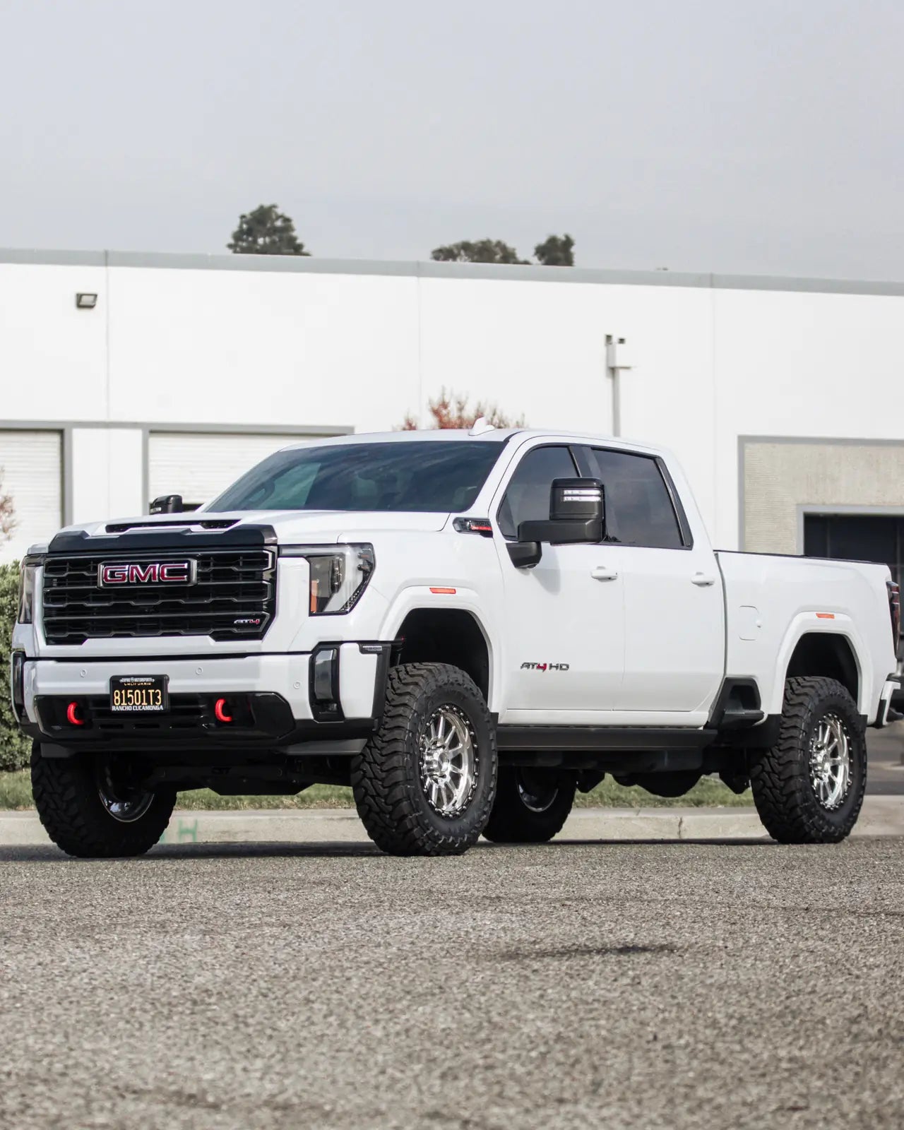 White GMC Sierra AT4HD from a distance at the front three-quarter, with polished wheels, mud tires and red tow hooks.