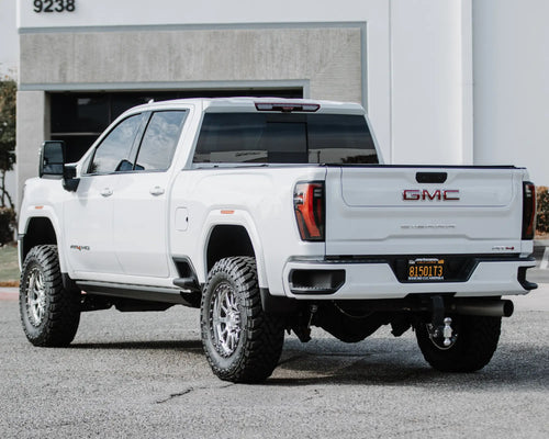 Rear three-quarter view of a lifted white GMC Sierra 2500 HD AT4 parked by a building