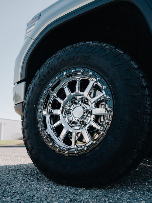 Close-up of a chrome multi-spoke wheel with an all-terrain tire on a silver GMC Sierra Denali