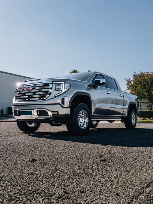 Front three-quarter view of a lifted silver GMC Sierra Denali with chrome wheels and all-terrain tires