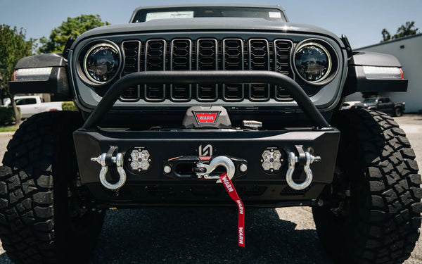 Close-up of a gray Jeep Wrangler with a steel front bumper, WARN winch, recovery shackles, and auxiliary lights
