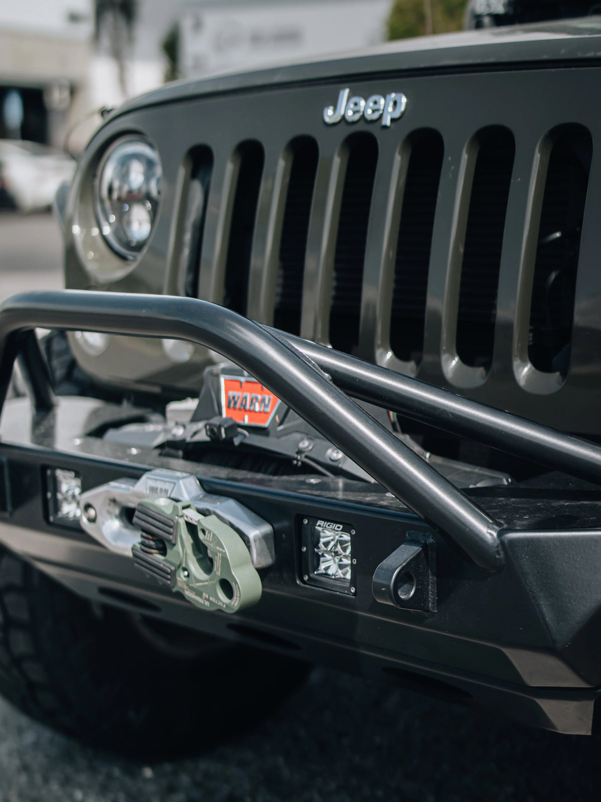 Close-up of a Jeep equipped with a steel off-road bumper, WARN winch, recovery hook, and RIGID LED lights.