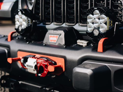 Close-up of a Warn winch with a red hook on a black Jeep Wrangler bumper between two Baja Designs LED lights