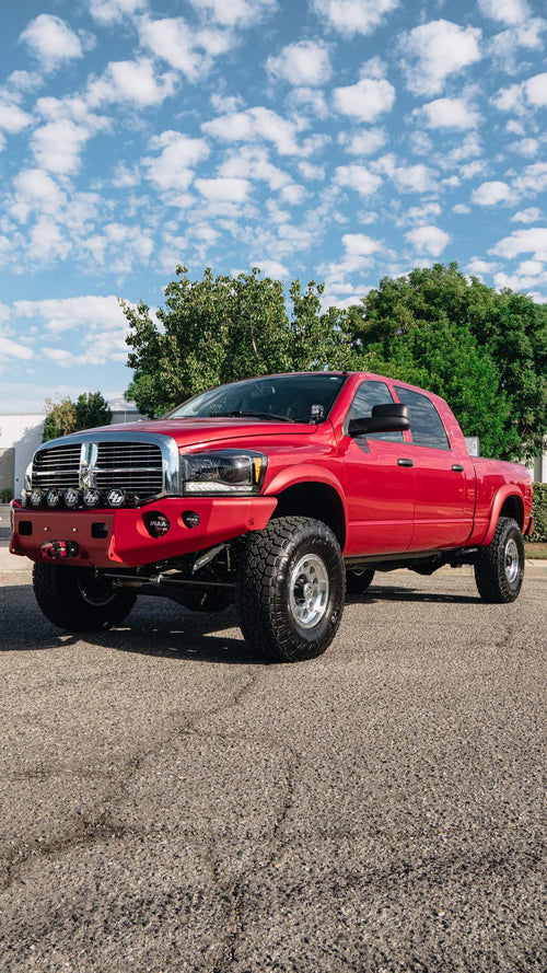 Front three-quarter view of a red Dodge Ram HD with a lighting and recovery build, parked on cracked asphalt