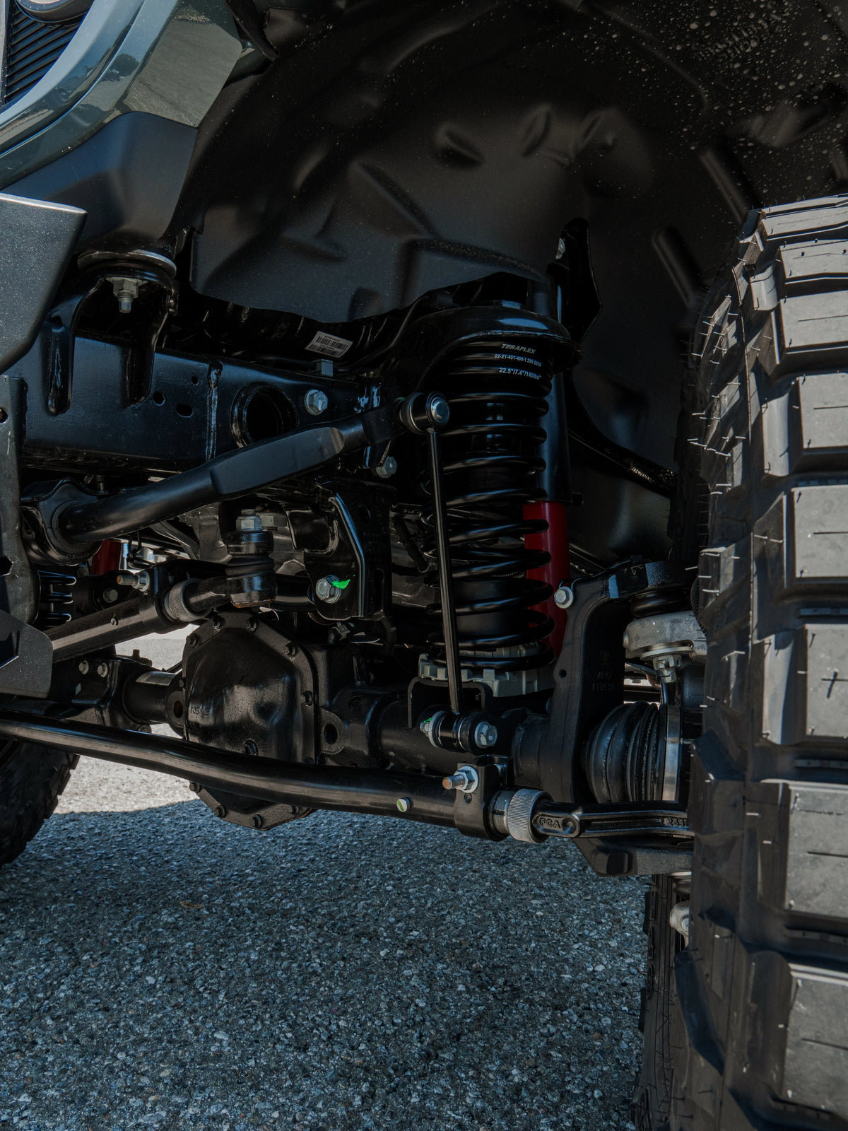 Close-up of a vehicle’s front suspension with a TeraFlex coil spring, shock absorber, steering components, and off-road tire.
